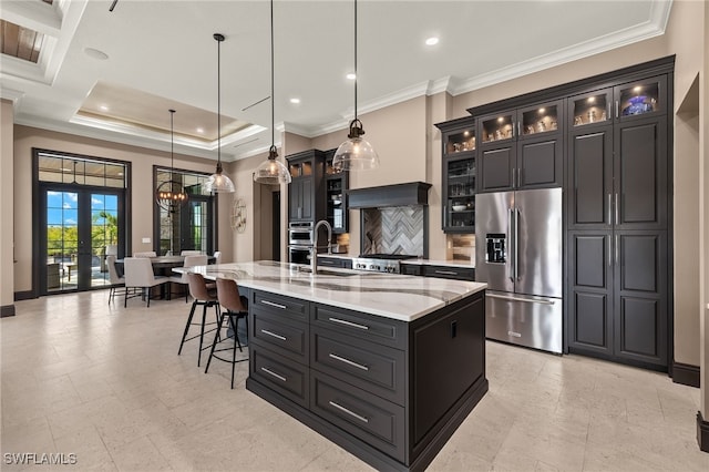 kitchen with ornamental molding, an island with sink, hanging light fixtures, light stone countertops, and high end fridge