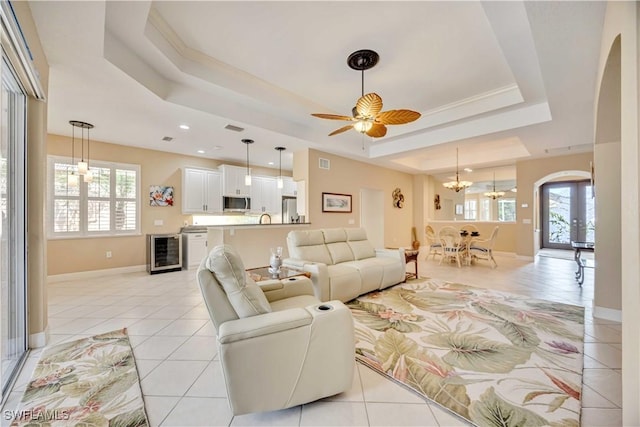 living room featuring french doors, wine cooler, a tray ceiling, light tile patterned floors, and ceiling fan with notable chandelier