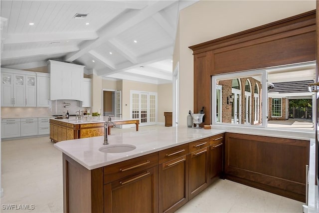 kitchen with light stone counters, sink, vaulted ceiling with beams, white cabinetry, and an island with sink