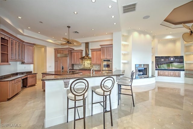 kitchen featuring wall chimney exhaust hood, a kitchen bar, built in refrigerator, a large island with sink, and dark stone counters