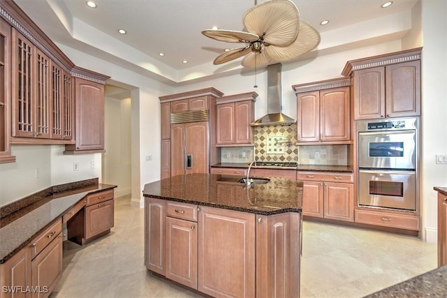 kitchen with an island with sink, wall chimney range hood, dark stone counters, appliances with stainless steel finishes, and ceiling fan