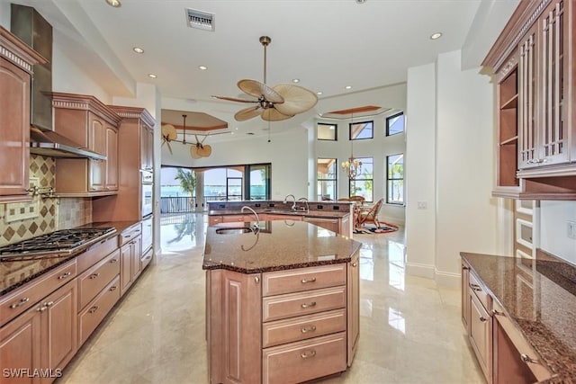 kitchen featuring dark stone counters, a kitchen island with sink, wall chimney range hood, and sink