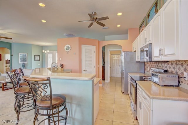 kitchen with light tile patterned floors, a breakfast bar area, white cabinetry, stainless steel appliances, and ceiling fan with notable chandelier