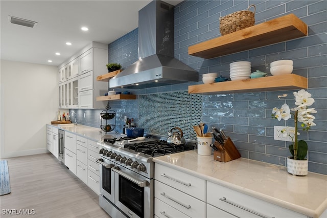 kitchen with tasteful backsplash, light stone counters, wall chimney range hood, range with two ovens, and white cabinetry