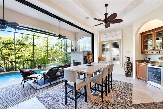 dining room featuring beverage cooler, ornamental molding, ceiling fan, a fireplace, and a tray ceiling