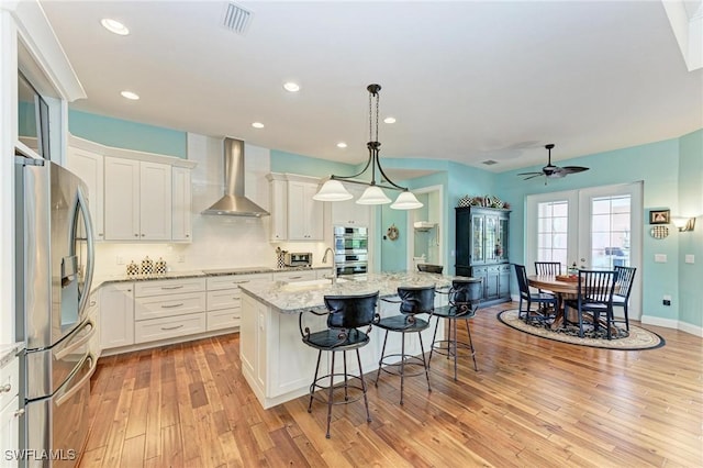 kitchen featuring hanging light fixtures, stainless steel appliances, wall chimney range hood, a kitchen island with sink, and white cabinets