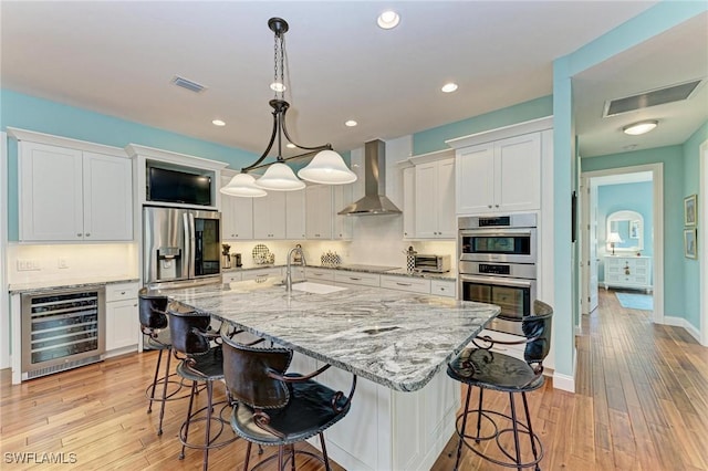 kitchen featuring white cabinets, wall chimney range hood, appliances with stainless steel finishes, decorative light fixtures, and beverage cooler