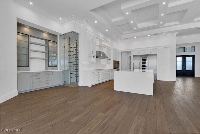 kitchen with french doors, wall chimney exhaust hood, cooktop, a center island with sink, and white cabinets