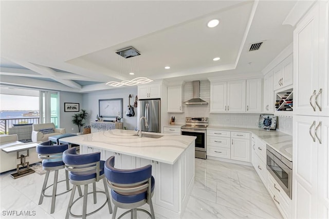 kitchen with a center island with sink, wall chimney exhaust hood, appliances with stainless steel finishes, decorative light fixtures, and a kitchen bar