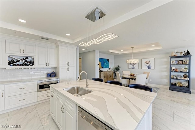 kitchen with sink, a raised ceiling, decorative light fixtures, a kitchen island with sink, and white cabinets