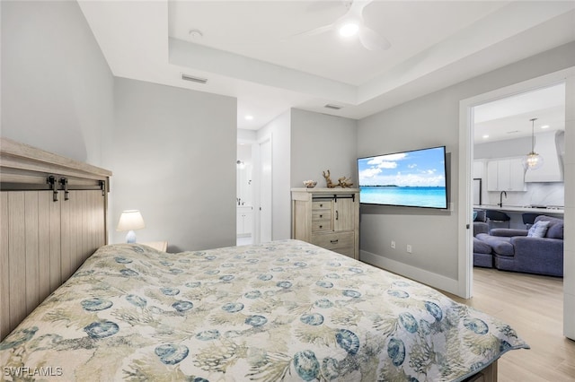 bedroom featuring ceiling fan, a barn door, ensuite bathroom, a raised ceiling, and light hardwood / wood-style flooring