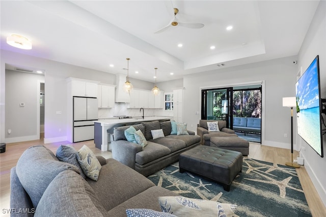 living room with a raised ceiling, ceiling fan, sink, and light hardwood / wood-style flooring