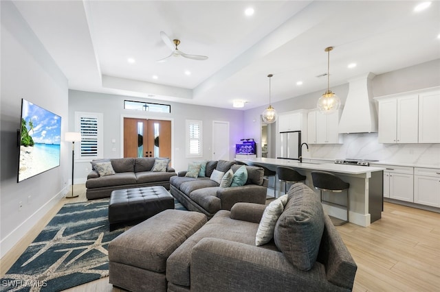 living room with ceiling fan, sink, a tray ceiling, light hardwood / wood-style flooring, and french doors