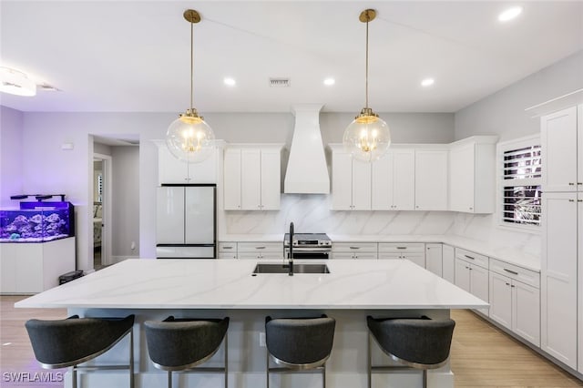 kitchen with white fridge, white cabinetry, custom exhaust hood, a center island with sink, and sink