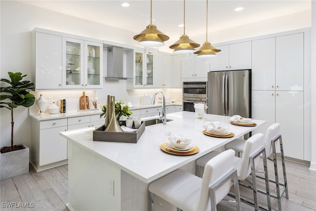 kitchen with white cabinetry, wall chimney exhaust hood, a center island with sink, appliances with stainless steel finishes, and light wood-type flooring