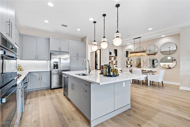 kitchen featuring appliances with stainless steel finishes, sink, a center island with sink, decorative light fixtures, and gray cabinets