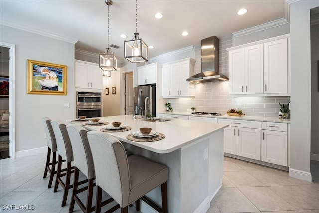 kitchen featuring white cabinets, appliances with stainless steel finishes, decorative light fixtures, wall chimney range hood, and a center island with sink