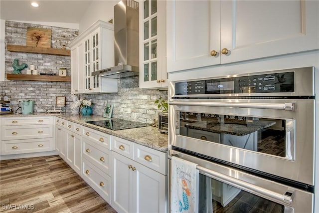 kitchen featuring tasteful backsplash, light stone counters, wall chimney exhaust hood, double oven, and white cabinets