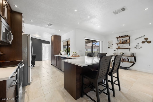 kitchen with a breakfast bar, dark brown cabinets, a textured ceiling, kitchen peninsula, and stainless steel appliances