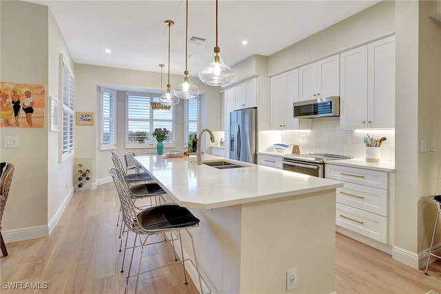 kitchen with sink, appliances with stainless steel finishes, a kitchen island with sink, white cabinets, and decorative light fixtures