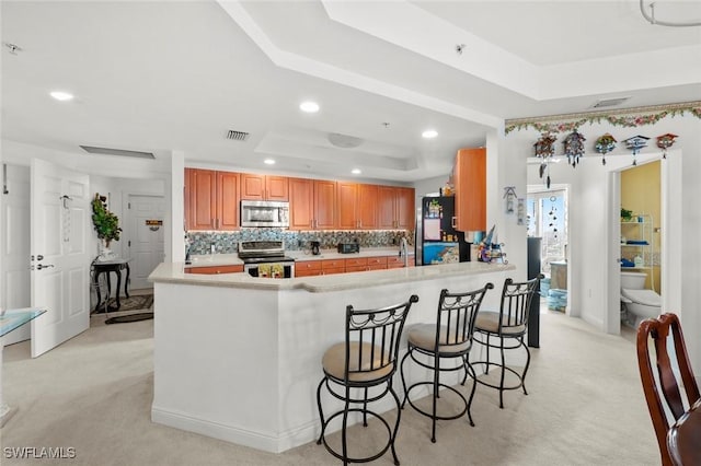 kitchen featuring a raised ceiling, decorative backsplash, appliances with stainless steel finishes, light colored carpet, and kitchen peninsula