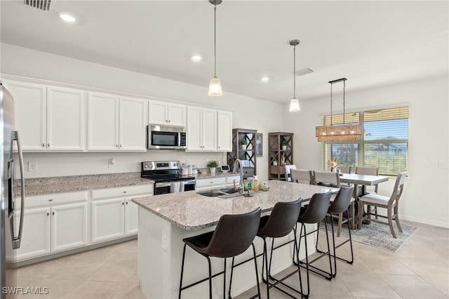 kitchen with light stone countertops, appliances with stainless steel finishes, a kitchen island with sink, sink, and decorative light fixtures