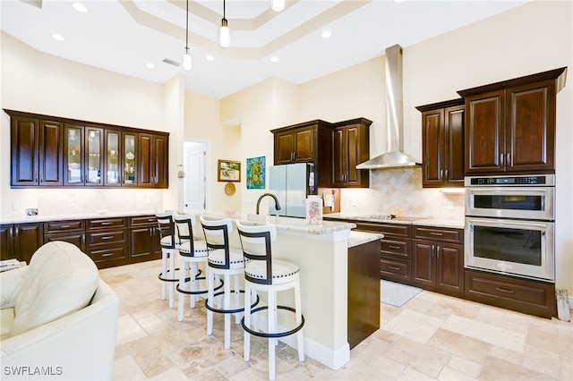 kitchen with refrigerator, wall chimney range hood, a towering ceiling, an island with sink, and double oven