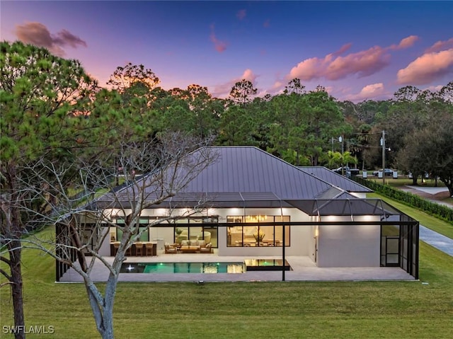 back house at dusk featuring a patio area, glass enclosure, and a lawn