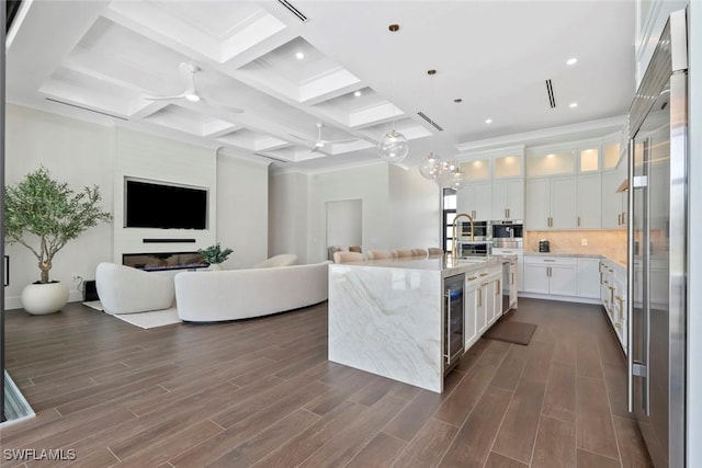 kitchen with white cabinetry, hanging light fixtures, coffered ceiling, light stone countertops, and a center island with sink