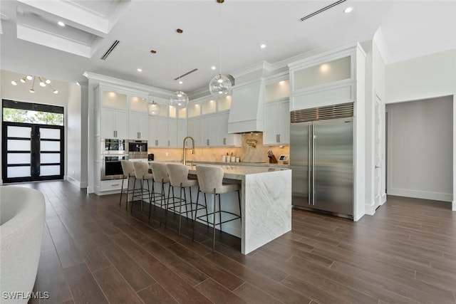 kitchen with built in refrigerator, light stone countertops, custom range hood, white cabinets, and a center island with sink