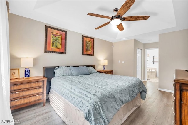 bedroom with ceiling fan, light hardwood / wood-style floors, a tray ceiling, and ensuite bath