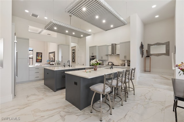 kitchen with a high ceiling, a kitchen island with sink, wall chimney exhaust hood, and gray cabinetry