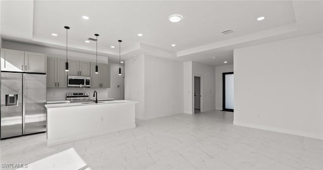 kitchen with a tray ceiling, stainless steel appliances, a kitchen island with sink, decorative light fixtures, and gray cabinets