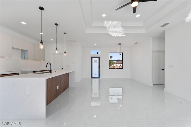 kitchen with sink, hanging light fixtures, a raised ceiling, a kitchen island with sink, and white cabinets