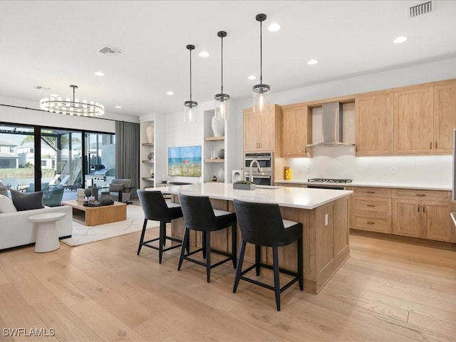 kitchen with wall chimney range hood, a large island with sink, pendant lighting, light wood-type flooring, and appliances with stainless steel finishes