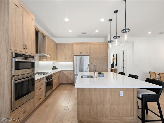 kitchen featuring wall chimney exhaust hood, a breakfast bar, a kitchen island with sink, light brown cabinets, and decorative light fixtures