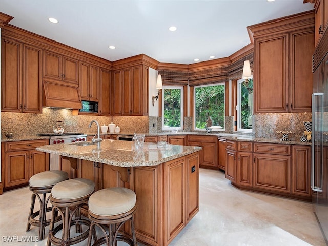 kitchen with built in appliances, light stone countertops, premium range hood, sink, and an island with sink