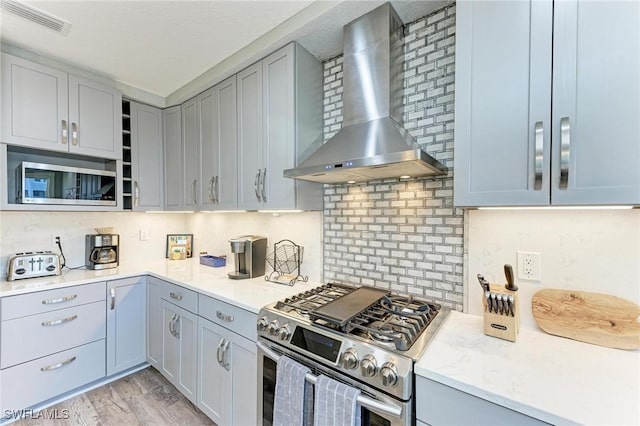 kitchen featuring wall chimney range hood, light stone countertops, light wood-type flooring, appliances with stainless steel finishes, and tasteful backsplash