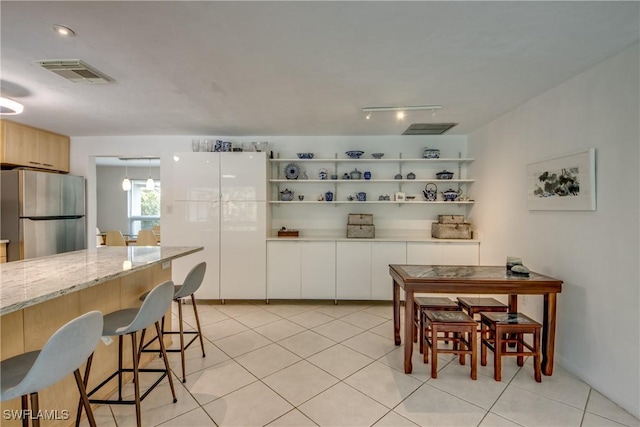 kitchen featuring stainless steel refrigerator, white cabinetry, a kitchen bar, light tile patterned floors, and light stone countertops