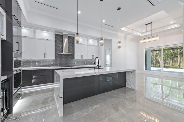 kitchen with sink, wall chimney range hood, a raised ceiling, a spacious island, and white cabinets