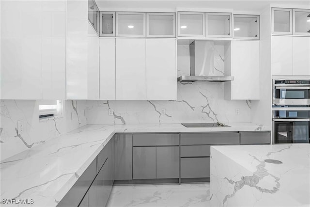 kitchen with gray cabinetry, black electric stovetop, wall chimney exhaust hood, light stone counters, and stainless steel double oven