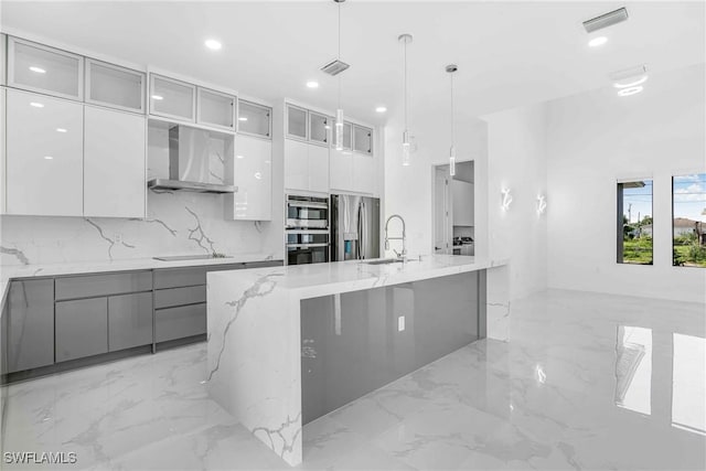 kitchen with light stone countertops, wall chimney exhaust hood, hanging light fixtures, stainless steel appliances, and a center island with sink