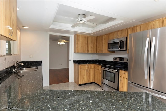 kitchen featuring ceiling fan, a raised ceiling, sink, appliances with stainless steel finishes, and dark stone counters