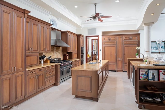 kitchen with range with two ovens, wall chimney range hood, decorative backsplash, sink, and a kitchen island with sink