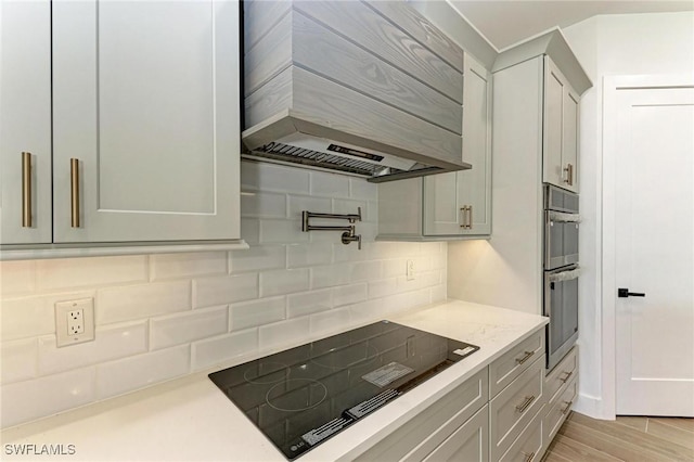 kitchen featuring wall chimney range hood, decorative backsplash, light stone countertops, light wood-type flooring, and black electric cooktop