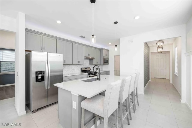 kitchen with gray cabinetry, stainless steel appliances, hanging light fixtures, and an island with sink