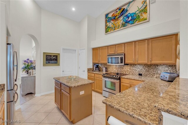 kitchen with light stone countertops, a high ceiling, a kitchen island, light tile patterned floors, and appliances with stainless steel finishes