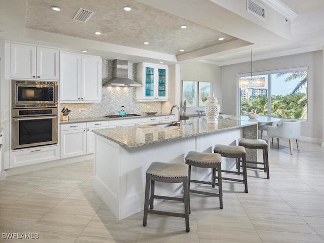 kitchen featuring stainless steel appliances, wall chimney range hood, a raised ceiling, a center island with sink, and white cabinets