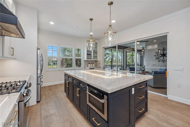 kitchen with wall chimney range hood, hanging light fixtures, light stone countertops, appliances with stainless steel finishes, and a kitchen island