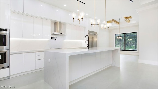 kitchen with white cabinets, wall chimney exhaust hood, stainless steel appliances, hanging light fixtures, and a center island with sink